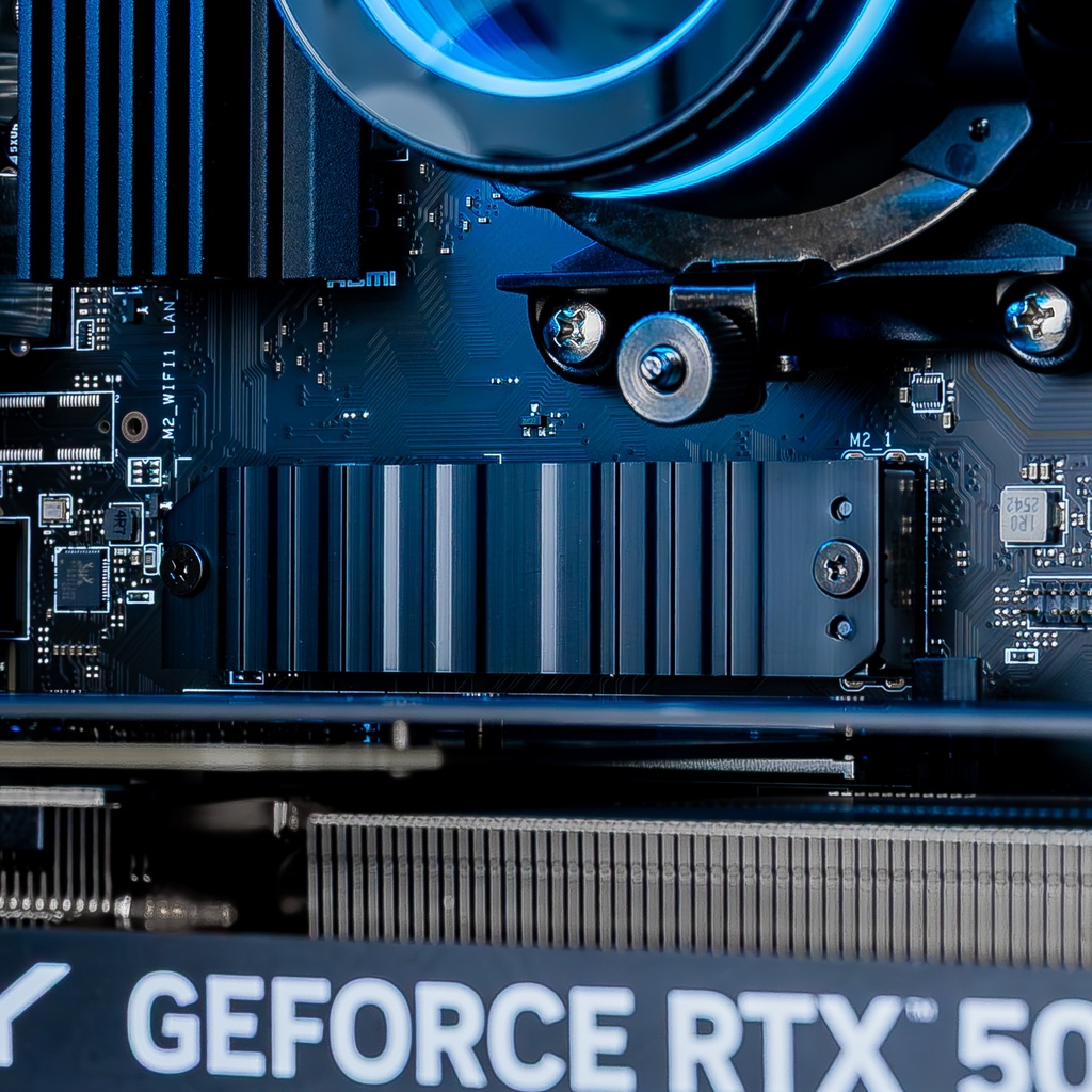 Gros plan sur le dissipateur thermique noir du SSD NVMe situé entre le waterblock AZZA éclairé en bleu et la carte graphique PNY GeForce RTX 5060 Ti du PC Gamer Savannah